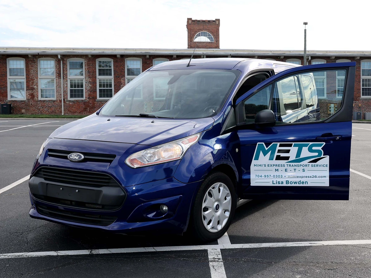 The navy-blue Mimi's Express Transport Service (METS) Ford Transit Connect van, door open to show the logo, phone number, and Lisa Bowden's name, parked outside a historic Charlotte mill building